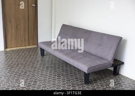Ein graues Stoffsofa-Bett auf gemustertem Fliesenboden in einem minimalen Flur mit Holztür und weißer Wand, ruhige moderne Innenszene mit sanftem natürlichem Licht Stockfoto