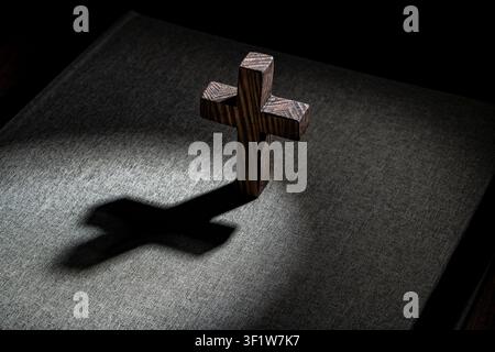 Holzernes christliches Kreuz wirft einen langen Schatten auf den Einband einer Bibel, Nahaufnahme. Kleines dunkles Holzkreuz steht auf einem stoffigen Bibeleinband und wirft Stockfoto