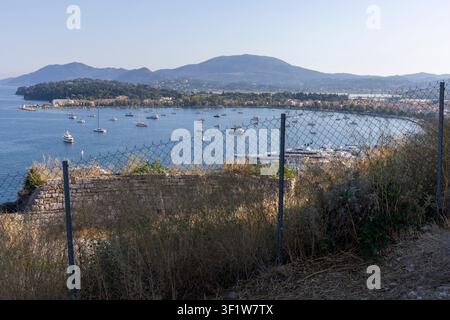Mit Blick auf die Bucht von Korfu-Stadt ankern zahlreiche Yachten und Segelboote in klaren Gewässern, mit fernen Bergen unter einem hellen Himmel Stockfoto