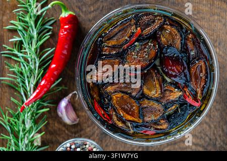 Sonnengetrocknete Pflaumen mit Knoblauch, grünem Rosmarin, rotem Chili, Olivenöl und Gewürzen in einem Glas auf einem Holztisch. Rustikaler Stil, Blick von oben, Nahaufnahme Stockfoto