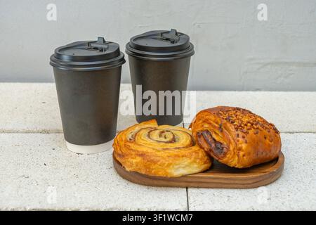 Zwei schwarze Einweg-Kaffeebecher mit einem Zimtschwirl und ein Schokoladen-Nussgebäck auf einem Holzbrett über einer Betonoberfläche. Stockfoto