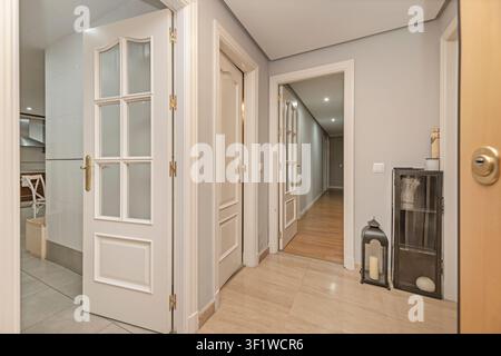 Flur mit weiß getäfelten Türen, goldenen Griffen, beigem Fliesenboden, einem dunklen Metallschrank, einer schwarzen Laterne und Blick in einen holzbeschichteten Korridor. Stockfoto