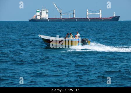 Ein kleines Boot mit örtlichen Passagieren navigiert durch die Gewässer der Straße von Hormuz in der Nähe der Insel Qeschm, mit einem großen Frachtschiff im Hintergrund. Stockfoto