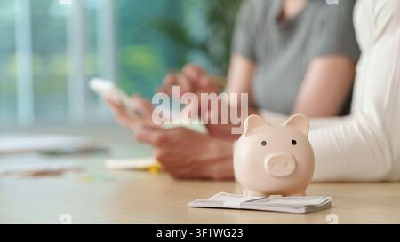 Rosa Sparschwein auf einem Stapel Dollar-Scheinen mit einem Paar, das im Hintergrund den Haushaltsplan plant Stockfoto