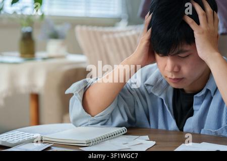 Gestresster asiatischer Mann, der den Kopf in den Händen hält, während er zu Hause Steuern berechnet Stockfoto
