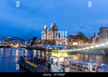 AMSTERDAM, NIEDERLANDE - 10. NOVEMBER 2025: Basilika des Heiligen Nikolaus bei Dämmerung über dem Kanal beleuchtet mit Touristenbooten in Amsterdam, Niederlande Stockfoto