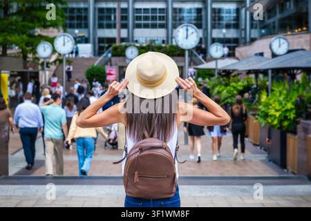 Eine Touristin mit Hut steht vor den berühmten Uhren in Canary Wharf, London, an einem sonnigen Sommertag Stockfoto