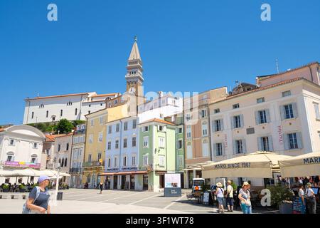 Piran, Slowenien. 26. Mai 2026. Ein sonniger Tartini-Platz ist von farbenfrohen Gebäuden und einem markanten Glockenturm gesäumt. Menschen sind zu sehen, die spazieren gehen und unter Sonnenschirmen im Freien speisen. Stockfoto