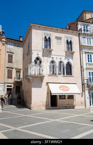 Piran, Slowenien. 26. Mai 2026. Ein blassrosa Gebäude mit dekorativen gotischen Fenstern und einem kleinen Balkon steht auf einem sonnigen Platz. Ein Vordach über einem Ladeneingang zeigt den Text Stockfoto
