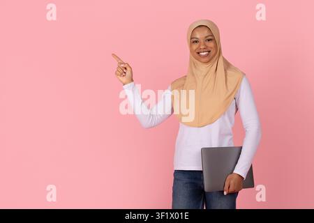 Schwarze muslimische Frau im Hijab hält einen Laptop und zeigt auf Kopierplatz Stockfoto