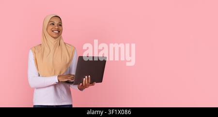 Lächelnde junge schwarze muslimische Frau in beigem Hijab, die an einem offenen Laptop arbeitet Stockfoto