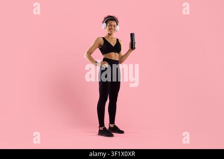 Vollständige Körperdarstellung einer glücklichen schwarzen Frau, die Kopfhörer trägt und eine Wasserflasche hält Stockfoto