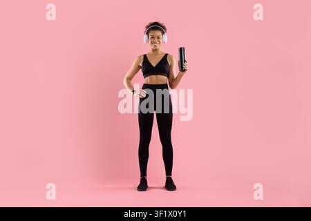 Fröhliche schwarze Frau in Sportkleidung, die kabellose Kopfhörer trägt und eine wiederverwendbare Flasche hält Stockfoto