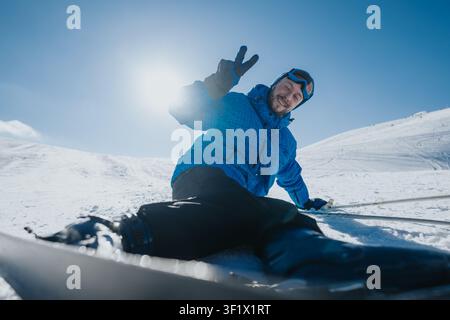 Mann auf Skipiste sitzt auf schneebedecktem Hang und zeigt Friedenszeichen, während er eine blaue Skijacke trägt Stockfoto