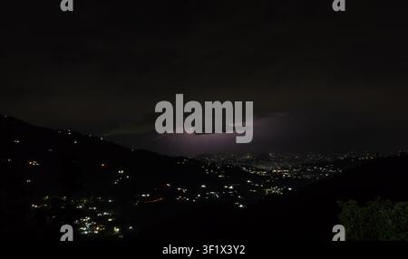 Eine dramatische Nachtlandschaft mit einem Blitzschlag, der dunkle, stimmungsvolle Gewitterwolken über einer weitläufigen Bergstadt beleuchtet. Stockfoto