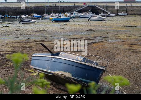 Kleines blaues Boot ruht auf schlammigem Ufer. Mehrere Boote liegen im Hintergrund am Dock. Grüne Pflanzen wachsen im Vordergrund. Das Wasser ist zurückgewichen und hat nassen Sand und Kieselstein freigelegt Stockfoto