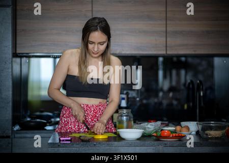 Junge Frau hackt glücklich Gemüse auf einem gelben Schneidebrett in einer modernen Küche. Hauskochen, gesunde Mahlzeitenzubereitung und tägliche Küchenroutine. Stockfoto