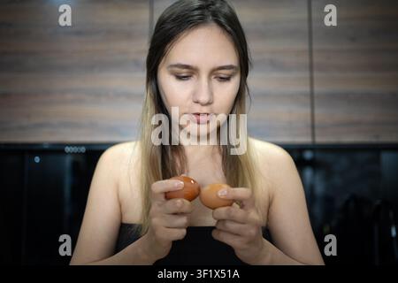 Junge Frau hält zwei braune Eier in ihren Händen, schaut mit konzentriertem Ausdruck nach unten. Kochvorbereitung, frische Zutaten und Morgenfrühstück Stockfoto