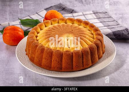 Frisch gebackener Bundt-Kuchen mit geriffeltem Design steht auf weißem Teller, begleitet von reifen Aprikosen und grünen Blättern auf karierter Stoffdecke, weiches natürliches Licht Stockfoto