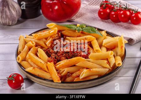 Leckere Penne-Pasta serviert mit reichhaltiger Fleischsauce und frischen Basilikumblättern auf rustikalem Teller. Kirschtomaten und Knoblauch im Hintergrund. Italienische Küche co Stockfoto