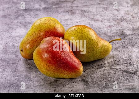 Drei reife Birnen mit gelber und roter Schale ruhen auf einer strukturierten grauen Steinoberfläche. Frische Bio-Früchte arrangiert in Dreieckkomposition mit natürlichem Li Stockfoto