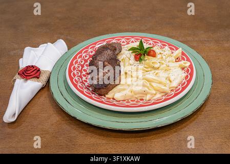 Hochwinkelaufnahme mit Steak und Penne-Pasta. Fokus auf cremige Textur, farbenfrohes Geschirr und Holzoberfläche. Ideal für Restaurantkonzept Stockfoto