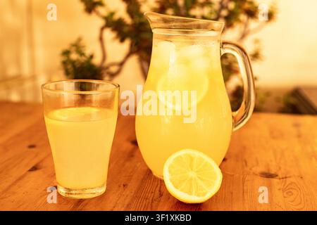 Ein Glaskrug und ein Tumbler mit frischer Limonade stehen auf einem Holztisch, garniert mit Zitronenscheiben, in goldenem Licht gebadet, ein erfrischendes Sommergetränk Stockfoto