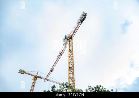 Niedrigwinklige Ansicht von zwei gelben Kranen auf der Baustelle unter einem blassblauen Himmel mit vereinzelten Wolken. Grünes Laub am unteren Rand setzt Kontrast Stockfoto