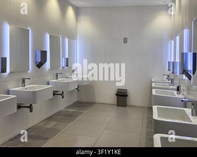 Ein sauberes, modernes öffentliches Toiletteninnere mit minimalistischen weißen Keramikwaschbecken, beleuchteten Spiegeln und professionellem Design in einem gewerblichen Büro Stockfoto