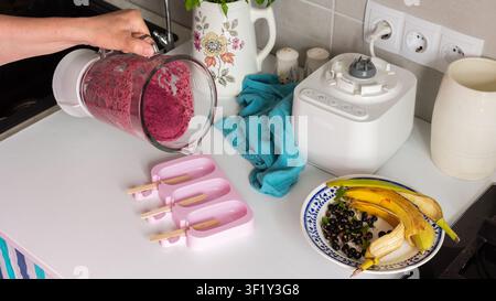 Eine Frau gießt eine Mischung aus Johannisbeere und Banane hausgemachtes Eis in rosa Formen in einer Küche. Es liegen Bananen und Beeren auf dem Tisch in der Nähe Stockfoto