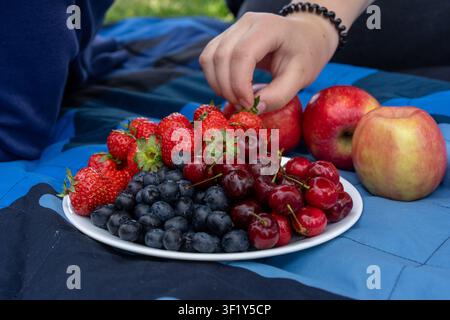 Eine Nahaufnahme im Querformat eines weißen Tellers gefüllt mit frischen reifen Erdbeeren, Blaubeeren und süßen Kirschen, daneben rote Äpfel auf einem bl Stockfoto