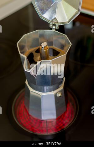 Offene Aluminium-Moka-Kanne mit dunklem Kaffee und Schaumbrühe auf einem glühend roten Elektroherd in einer Küche. Stockfoto