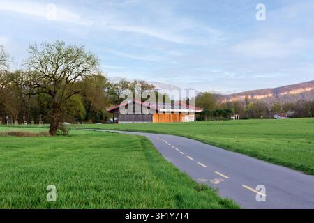 Ein gebogener gepflasterter Radweg führt durch grüne Felder zu einer Holzbarn mit Bergen im Hintergrund in der Nähe von Genf, Schweiz. Stockfoto