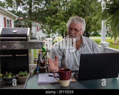 Ein Mann sitzt an einem Tisch und arbeitet an einem Laptop, während er einen Kaffee genießt. Er befindet sich draußen auf einer Veranda neben einem Grill. Es ist ein sonniger Nachmittag, und um ihn herum ist Grün sichtbar. Stockfoto