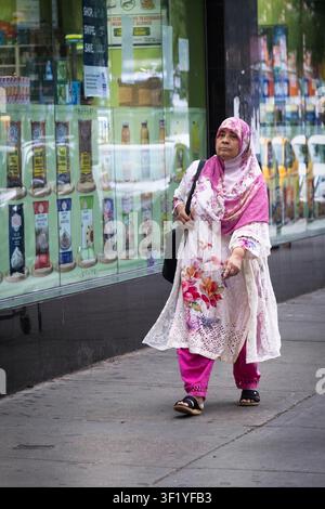 Eine bescheiden gekleidete muslimische Frau mit einem farbenfrohen Outfit und einem Hijab geht in der 74th Street im Viertel Little Bangladesh von Queens, New York. Stockfoto