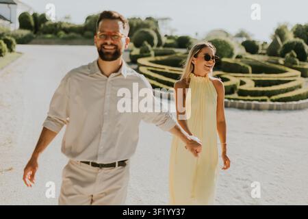 Ein Paar schlendert Hand in Hand durch einen atemberaubenden Garten, sonnt sich im Sonnenschein und lacht. Ihr Glück strahlt in der ruhigen Umgebung. Stockfoto