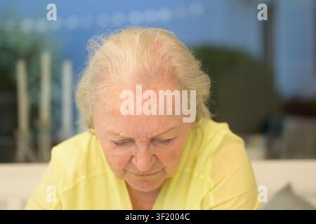 Eine nachdenkliche Seniorin schaut konzentriert nach unten, während sie in einer ruhigen und entspannten häuslichen Umgebung drinnen sitzt. Stockfoto