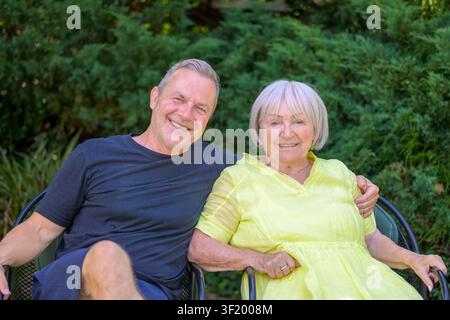 Lächelnder erwachsener Sohn und ältere Mutter genießen einen entspannten Moment zusammen im Garten und blicken in die Kamera. Stockfoto