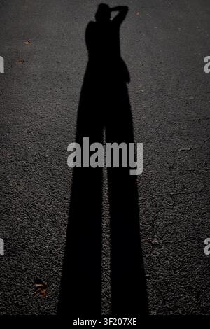 Silhouette einer Person auf Stelzen steht groß. Der Schatten erstreckt sich weit über den Asphalt. Kopf nach hinten geneigt, Arme über dem Kopf erhoben. Dramatische Beleuchtung erzeugt kräftige Stockfoto