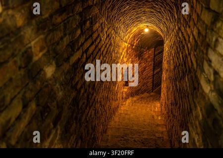 Tunnel mit Ziegelwänden und Treppen, die in einen dunklen, beleuchteten unterirdischen Raum hinabführen Stockfoto