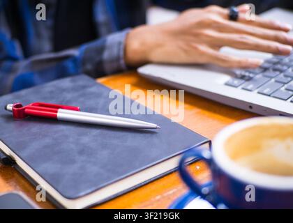 Mann arbeitet an seinem Laptop in einem Café mit Nahaufnahmen einer blauen Keramik-Kaffeetasse, eines Notizbuchs mit einem Stift und Details eines Remote-Arbeitsplatz-Setups Stockfoto