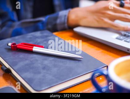 Mann arbeitet an seinem Laptop in einem Café, mit Nahaufnahmen einer blauen Keramik-Kaffeetasse, eines Notizbuchs mit einem Stift und Details eines Remote-Arbeitsplatz-Setups Stockfoto