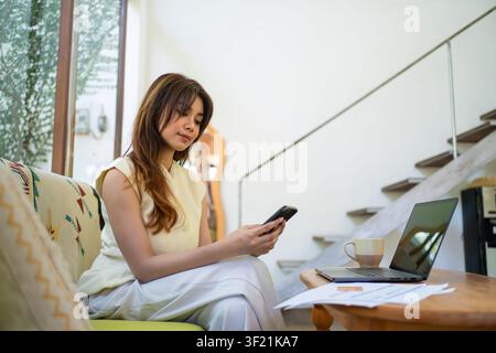 eine Frau, die ein Smartphone neben einem Laptop und Dokumenten verwendet, was Multitasking, Fernarbeit und digitale Kommunikation im modernen Arbeitsplatz darstellt. Stockfoto