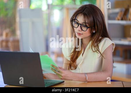 Asiatische Geschäftsfrau mit Brille, die Papierkram analysiert, während sie im Büro an einem Laptop arbeitet. Stockfoto