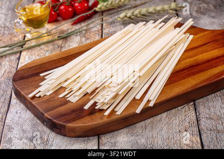 Ungekochte Udon-Nudeln auf rustikalem Holzbrett arrangiert. Hintergrund enthält Kirschtomaten, Olivenöl, Weizenhalme. Warme natürliche Beleuchtung hebt hervor Stockfoto