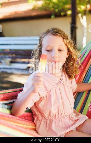 Niedliches Kind mit Eis am Stiel, das in einem gestreiften Hammock im sonnigen Hinterhof entspannt Stockfoto