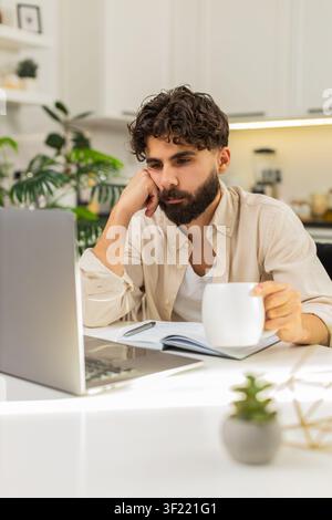 Ein indischer Mann zu Hause arbeitet am Laptop und fühlt sich nach der Arbeit sehr müde. Er hält Kopf mit der Hand in die Nähe einer Tasse Kaffee und Dokumente. Arabianer, der am Tisch sitzt, schaut auf die Bildschirme, fühlt sich schwer, dass Burnout Ruhe will. Stockfoto