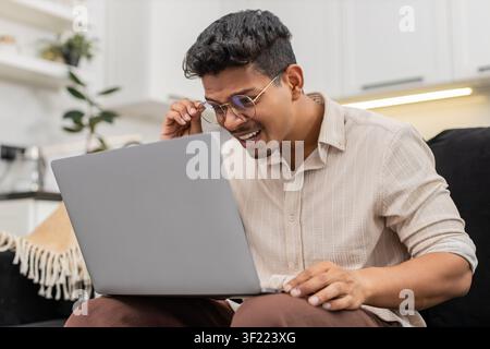Ein indischer Mann zu Hause hat Schwierigkeiten, winzige Texte auf dem Laptop zu lesen, zieht dann eine Brille auf und sieht klare Sicht. Arabian Guy on Sofa Lächeln passt den Rahmen an, liest Bildschirmlinien ohne Belastung und setzt die Arbeit fort. Stockfoto