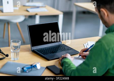 Unerkennbarer Mann, der Notizen schreibt, während er einen Laptop an einem modernen Bürotisch verwendet, Produktivitätskonzept Stockfoto