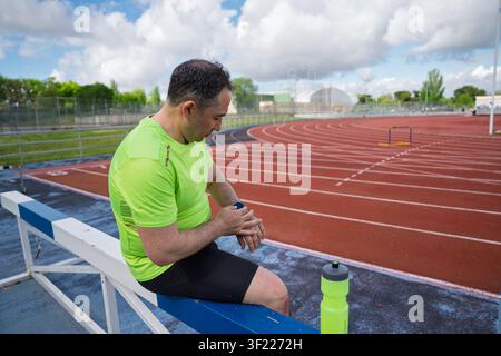 Mann ruht sich auf einem Hindernis auf einem Sportstadion aus und nutzt sein tragbares Fitnessgerät Stockfoto